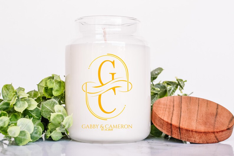 Puede incluir: Una vela blanca en un tarro de cristal con tapa de madera. La vela tiene un monograma dorado y el texto "Gabby & Cameron" con una fecha. El follaje verde est&aacute; en el fondo.