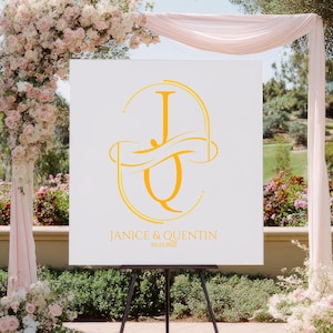 Puede incluir: Un letrero de bienvenida de boda blanco con un monograma dorado de las letras "J" y "Q" se muestra en un caballete negro. El letrero tambi&eacute;n incluye los nombres "Janice & Quentin" y la fecha de la boda. El fondo presenta un arco floral.
