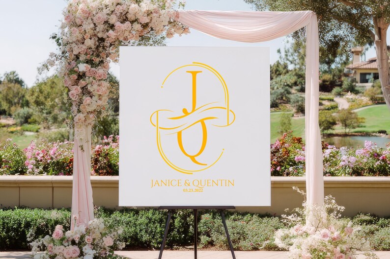 Puede incluir: Un letrero de bienvenida de boda blanco con un monograma dorado de las letras "J" y "Q" se muestra en un caballete negro. El letrero tambi&eacute;n incluye los nombres "Janice & Quentin" y la fecha de la boda. El fondo presenta un arco floral.