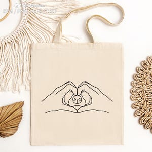 Puede incluir: Una bolsa de tela blanca con un dibujo de l&iacute;nea negra de dos manos haciendo un coraz&oacute;n con la nariz de un perro en el centro.