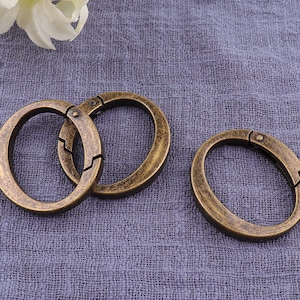 May include: Three antique gold-toned metal key rings with a spring-loaded clasp. The key rings are lying on a textured, light purple fabric surface.