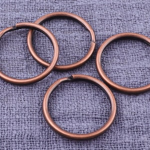 May include: Four copper-colored key rings, each with a circular shape, are arranged on a textured purple fabric background.