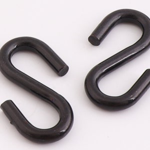Könnte beinhalten: Zwei schwarze Metall-S-Haken. Die Haken bestehen aus einem soliden, dunklen Material und haben eine glatte, leicht reflektierende Oberfläche. Das S-förmige Design ermöglicht das einfache Aufhängen und Verbinden von Gegenständen.