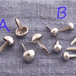 May include: A close-up of silver metal rivets with a domed head and a straight shank. The rivets are arranged on a gray fabric background. The letters A and B are written in blue on the fabric.