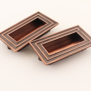 May include: Two rectangular, copper-colored cabinet pulls with a recessed center. The pulls have a layered, multi-dimensional design.