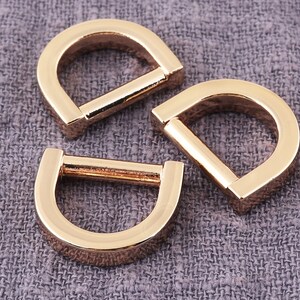 Puede incluir: Tres anillos en D de metal de color dorado. Los anillos son lisos y brillantes. Están dispuestos en forma de triángulo sobre un fondo de tela gris.
