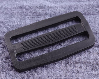 May include: A black plastic slider buckle for webbing or straps. The buckle has a rectangular shape with a smooth, flat surface and a raised, textured center section.