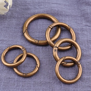 Pode incluir: Cinco chaveiros de metal dourado antigo com fecho de mola. Os chaveiros são de tamanhos diferentes e estão dispostos em semicírculo sobre uma superfície de tecido cinzento texturizado.