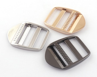 May include: Three metal slider buckles in silver, gold, and black. Each buckle has a rectangular shape with rounded edges and three parallel slots for a strap.