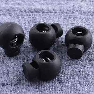 May include: Four black cord locks with a spring mechanism. The cord locks are spherical with a cylindrical base and a hole for the cord. The background is a textured, light blue fabric.
