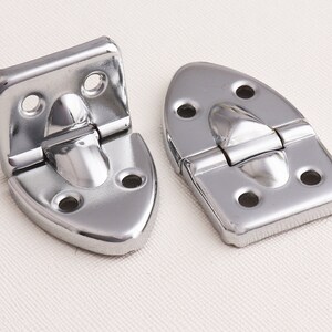 May include: Two silver-toned metal hinges with a rounded, slightly pointed shape. The hinges are closed and have a polished finish.