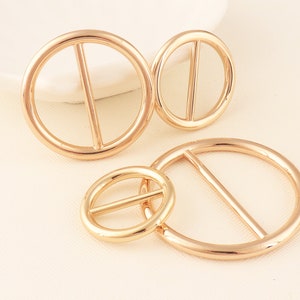 May include: Four gold-colored metal belt buckles in varying sizes. Each buckle has a circular shape with a single bar across the center. The buckles are arranged on a light-colored surface.
