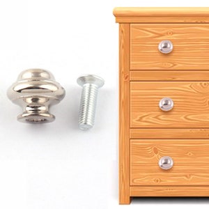 Può includere: Pomo per armadio argentato con una parte superiore rotonda leggermente bombata e un anello decorativo attorno alla base. Il pomo è fissato a una vite argentata.