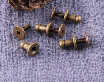 May include: Five antique gold-toned metal screw-on knobs with a round head and a small, round knob on top. The knobs are on a gray fabric surface.