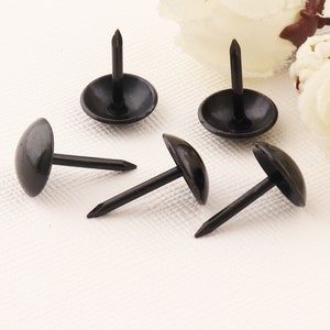 Peut inclure: Cinq clous en métal noir avec des têtes rondes et des pointes acérées. Les clous sont disposés en demi-cercle sur une surface blanche.