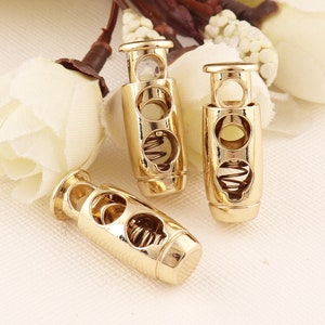 May include: Three gold-toned toggle cord locks with a spring mechanism. Each lock has two round holes and a spring inside. The locks are on a white surface.