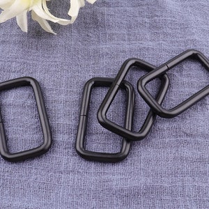 Peut inclure: Cinq anneaux en D en métal noir, également appelés anneaux en D, sont disposés sur une surface en tissu gris. Les anneaux sont de forme rectangulaire avec des coins arrondis.