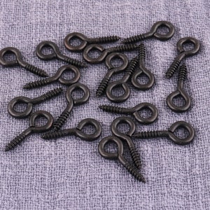 May include: A close-up of a group of black metal eyelet screws. The screws have a loop at the top and a pointed end. They are scattered on a gray fabric surface.