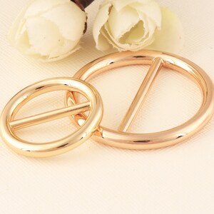 May include: Two gold-colored metal belt buckles. One is oval-shaped, and the other is a larger circle with a bar across the center. The buckles are shiny and reflect light, suggesting a polished finish. The background includes white flowers.