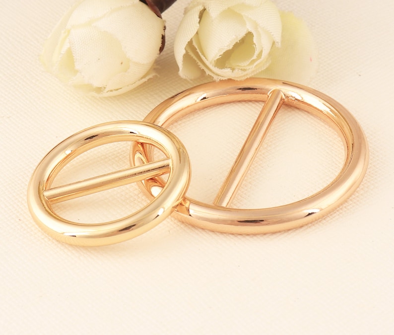 May include: Two gold-colored metal belt buckles. One is oval-shaped, and the other is a larger circle with a bar across the center. The buckles are shiny and reflect light, suggesting a polished finish. The background includes white flowers.