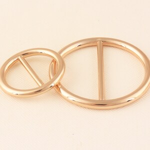 May include: Two gold-colored metal belt buckles. One is a smaller circle, and the other is a larger oval shape. Each buckle has a single bar across the center. The buckles are linked together.