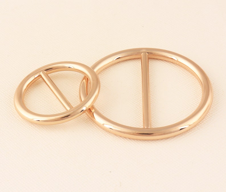 May include: Two gold-colored metal belt buckles. One is a smaller circle, and the other is a larger oval shape. Each buckle has a single bar across the center. The buckles are linked together.