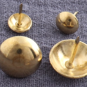 Puede incluir: Cuatro tachuelas de metal de color dorado con cabeza redonda y clavo puntiagudo. Las tachuelas están dispuestas en forma de cuadrado sobre una superficie de tela gris.