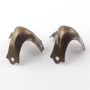 Op de afbeelding: Twee antieke gouden hoekbeschermers met een decoratief ontwerp. De beschermers zijn gemaakt van metaal en hebben een verweerde afwerking.