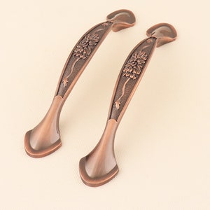 May include: Two antique copper cabinet pulls with a floral design. The pulls are curved and have a decorative floral pattern.