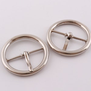 Könnte beinhalten: Zwei silberne Metall-Gürtelschließen. Jede Schließe hat eine runde Form mit einem Metallbügel in der Mitte.