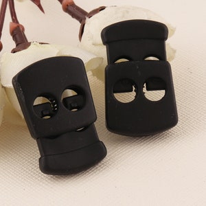 May include: Two black plastic cord locks with two holes each. The cord locks are designed to secure cords or straps.