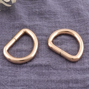 May include: Two gold-colored D-rings, each with a flat, straight section and a rounded, D-shaped portion. The rings are made of a shiny metal and are displayed on a textured, purple-gray fabric.