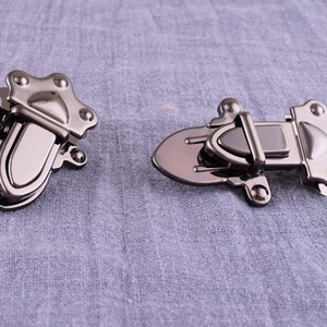 Könnte beinhalten: Zwei silberfarbene Metallverschlüsse für eine Tasche oder Geldbörse. Ein Verschluss ist ein einfacher Haken und der andere ist ein aufwendigeres Design mit einem Scharnierdeckel.