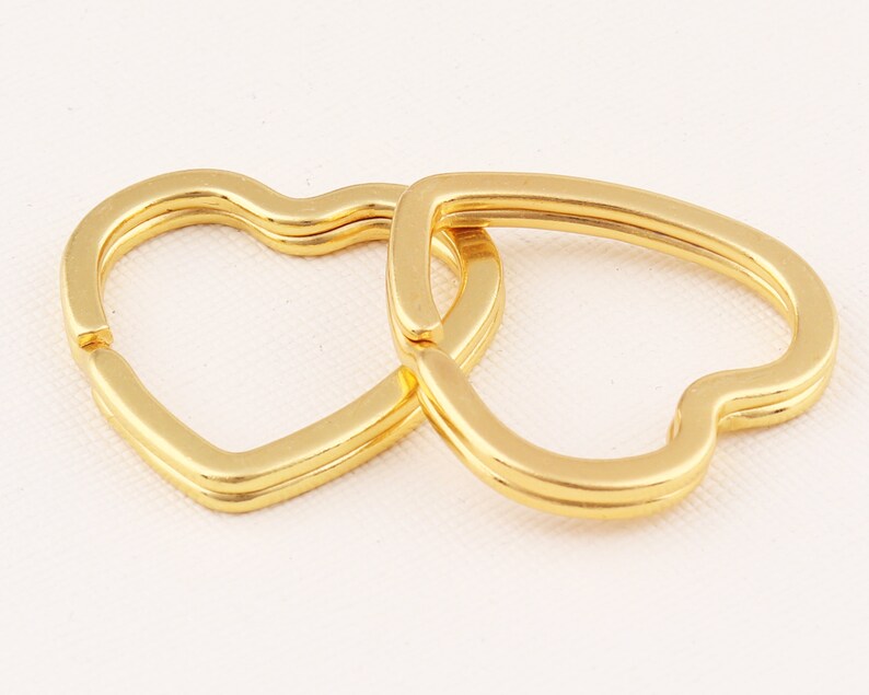 K&ouml;nnte beinhalten: Zwei goldene herzf&ouml;rmige Schl&uuml;sselanh&auml;nger, die miteinander verbunden sind. Die Schl&uuml;sselanh&auml;nger sind aus Metall und haben eine gl&auml;nzende Oberfl&auml;che.