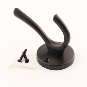 Puede incluir: Gancho de metal negro con base redonda y un solo gancho. El gancho se fija a la pared con dos tornillos y tacos de plástico.