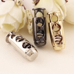 May include: Three metal cord stoppers in silver, black, and gold. Each stopper has a spring mechanism and two holes for threading cord.