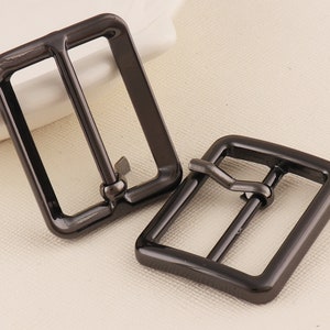 May include: Two black metal belt buckles. The buckles are rectangular with a smooth, shiny finish. The buckles have a simple design with a single bar that slides through the loop.