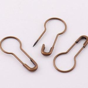 May include: Three antique gold safety pins. The pins are all the same size and shape, with a rounded head and a sharp point. They are made of metal and have a slightly tarnished finish.