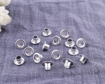 May include: Silver metal eyelets with a small hole in the center, scattered on a gray fabric surface.