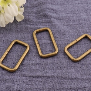 May include: Three gold-toned metal rectangular rings, also known as D-rings, are shown on a textured gray fabric surface. The rings are approximately 1 inch in length and 1/2 inch in width.