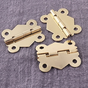 May include: Three gold-toned metal hinges with decorative details. Each hinge has a different shape and size.