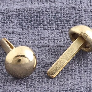 Puede incluir: Dos remaches de metal de color dorado con cabeza redonda y vástago recto. Los remaches están sobre un fondo de tela gris.