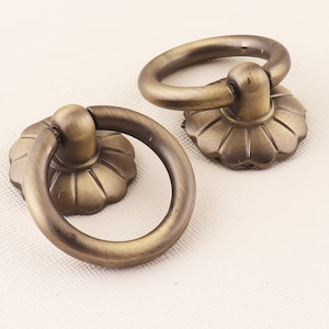 Op de afbeelding: Twee antieke messing ringgrepen met een decoratief bloemmotief. De grepen hebben een ronde ring en een decoratieve basis.