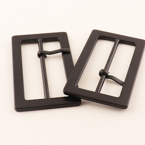 Puede incluir: Dos hebillas de cinturón de metal negro con forma rectangular y acabado liso.