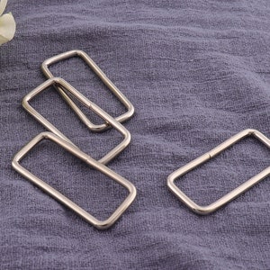 May include: Four silver-colored rectangular metal rings. The rings are evenly spaced on a textured, blue-gray fabric. The rings have rounded corners and a shiny finish. These rings are likely used for bag making or other craft projects.