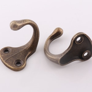 Könnte beinhalten: Zwei antike goldfarbene Metallhaken mit abgerundeter Hakenform und einer flachen Basis mit zwei Löchern zur Montage.