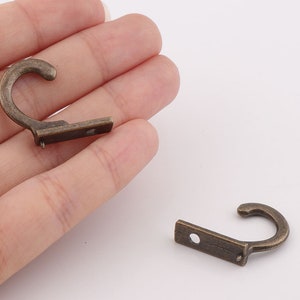 May include: Two antique gold-toned metal hooks with a flat base and a curved hook. The hooks are about 2 inches long.