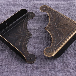 May include: Two antique gold corner brackets with an ornate design. The brackets are made of metal and have a distressed finish.