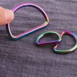 May include: Two iridescent metal D-rings, one larger than the other, are shown on a textured gray fabric background.
