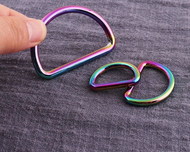 May include: Two iridescent metal D-rings, one larger than the other, are shown on a textured gray fabric background.